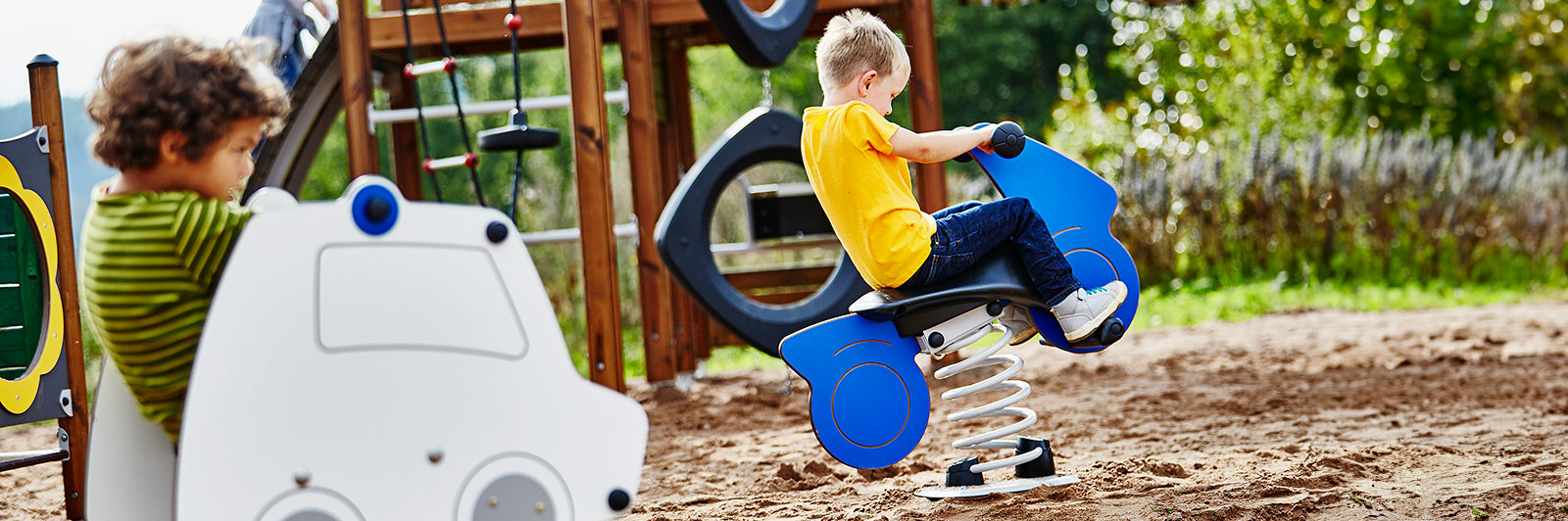 Two boys play on vehicle shaped springer toys at a playground, it looks like their are attempting to race each other.